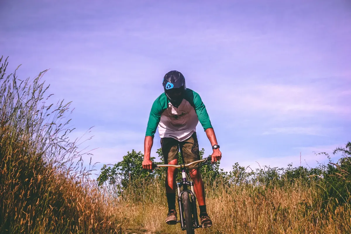A mountain biker rides through a grassy trail in Nelson, New Zealand, wearing a helmet and casual attire.
